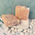 The soap bar has a soft, warm appearance with delicate swirls of peach, blush, and creamy ivory blending together throughout the surface. The marbling creates a gentle, cloud-like pattern that flows naturally through the bar. The top has a lightly textured, hand-shaped ridge that adds an artisanal look, while the sides reveal subtle layers of the peach and cream tones. The overall design feels smooth, warm, and elegant, with each swirl forming a unique pattern in the bar. 