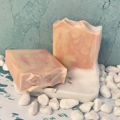 The soap bar has a soft, warm appearance with delicate swirls of peach, blush, and creamy ivory blending together throughout the surface. The marbling creates a gentle, cloud-like pattern that flows naturally through the bar. The top has a lightly textured, hand-shaped ridge that adds an artisanal look, while the sides reveal subtle layers of the peach and cream tones. The overall design feels smooth, warm, and elegant, with each swirl forming a unique pattern in the bar. 