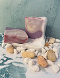 Two bars of soap with marbled light purple and white patterns and a bottom layer of purple with a pink line through the middle of the soap and topped with pink mica shimmer swirls.