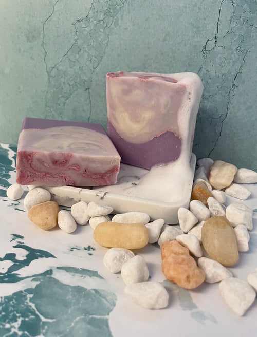 Two bars of soap with marbled light purple and white patterns and a bottom layer of purple with a pink line through the middle of the soap and topped with pink mica shimmer swirls.