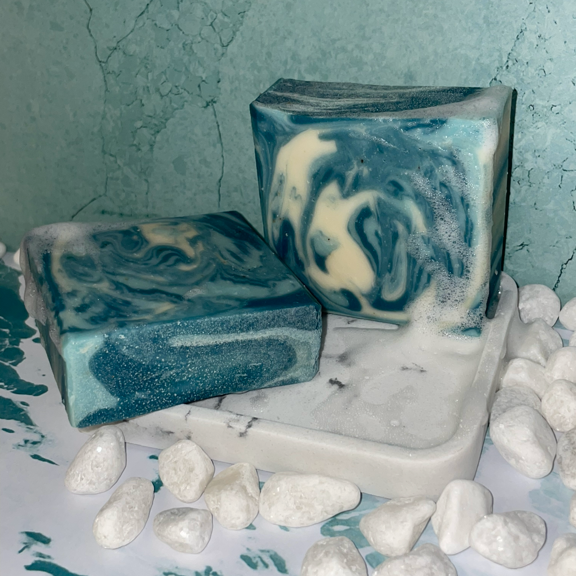 The soap bar features a striking swirl of deep teal, aqua, and creamy white tones. The colors flow together in a fluid, wave-like pattern, creating the look of ocean water moving through foam. The top has a gently textured, slightly curved surface, while the sides reveal layered ribbons of teal and pale blue with soft white marbling throughout. The design gives the bar a cool, aquatic feel, with each swirl forming a unique, natural pattern.