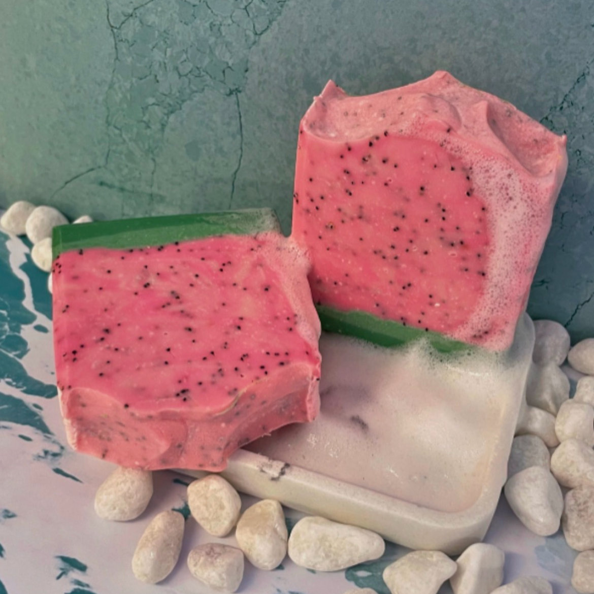 A bright pink soap bar with a small light green and dark green bottom layer to resemble a watermelon rind. Pink center layer is full of exfoliating poppy seeds to resemble watermelon seeds in a slice of watermelon. 