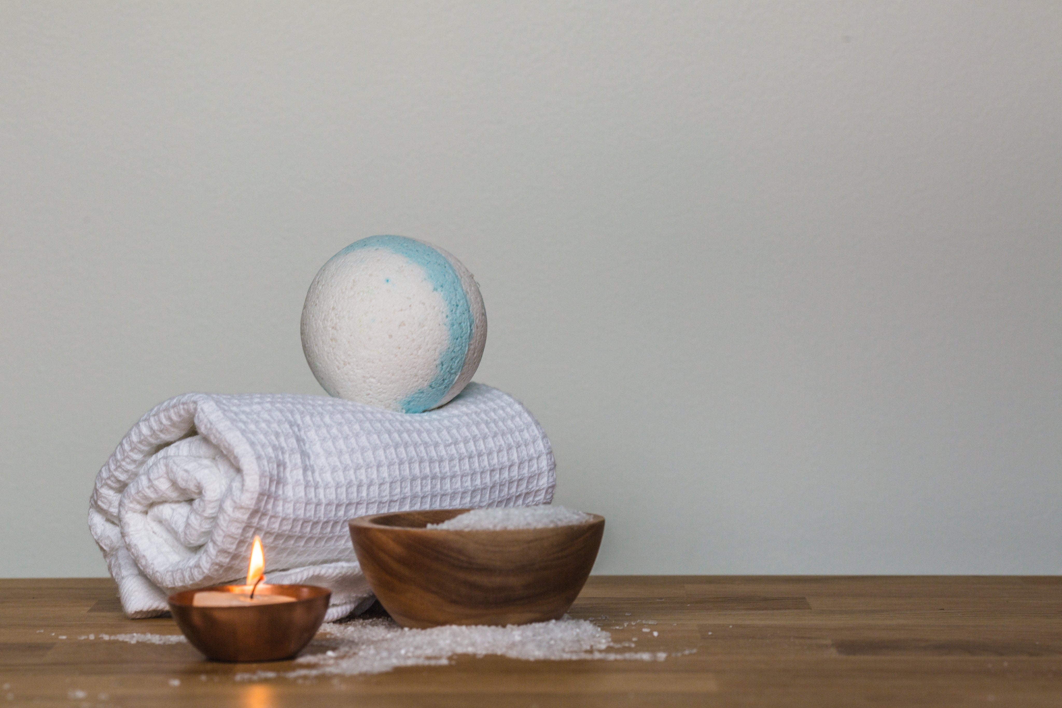 Spa still life with a rolled white towel topped by a bath bomb, a small lit candle, and a wooden bowl of bath salts on a wooden surface against a plain light background.