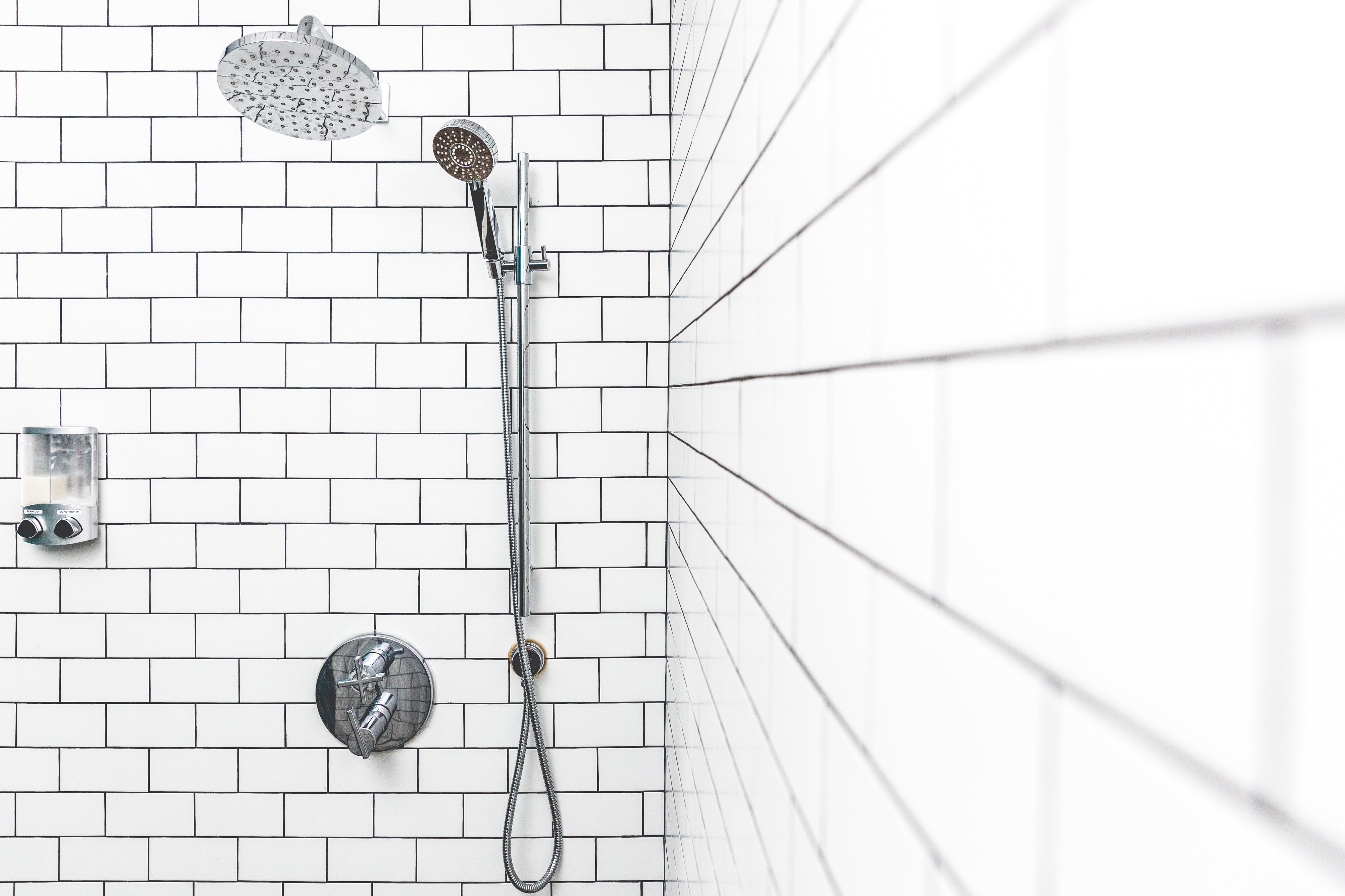 Wall-mounted shower with a large round rainfall showerhead and handheld sprayer on a vertical rail, set against white subway tile walls with gray grout lines.
